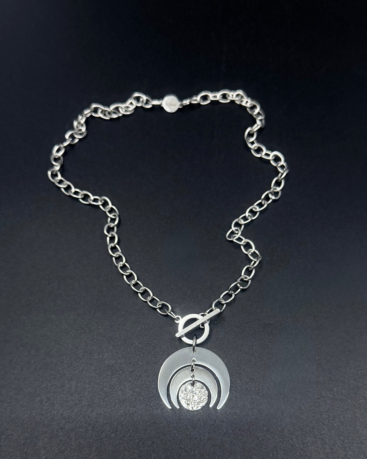 Silver Luna Phase necklace with crescent moon pendant and textured disc on a stainless steel curb chain with toggle clasp.