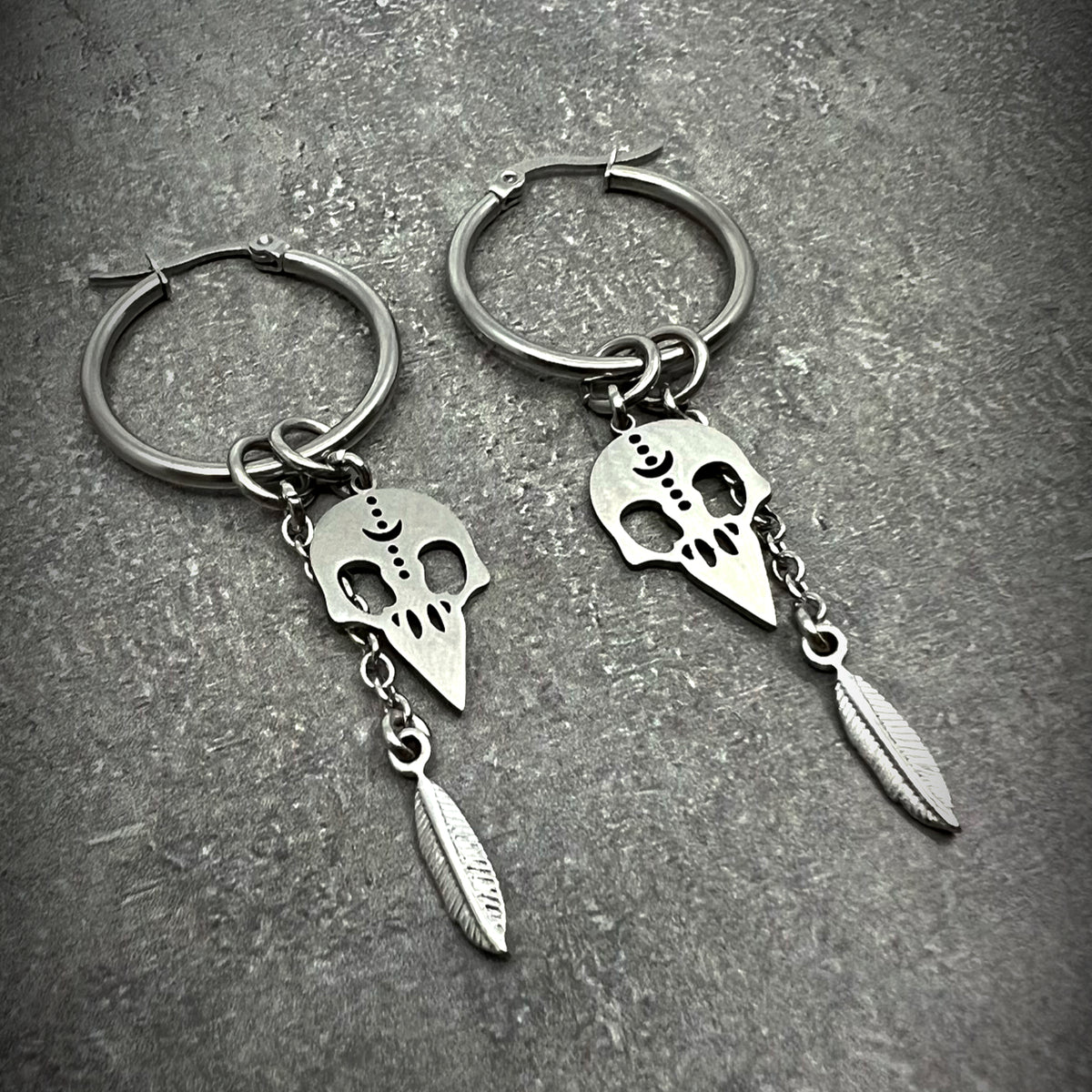 All-silver stainless steel hoop earrings featuring skull and feather charms, sculptural lightweight design — Lawless Jewellery.