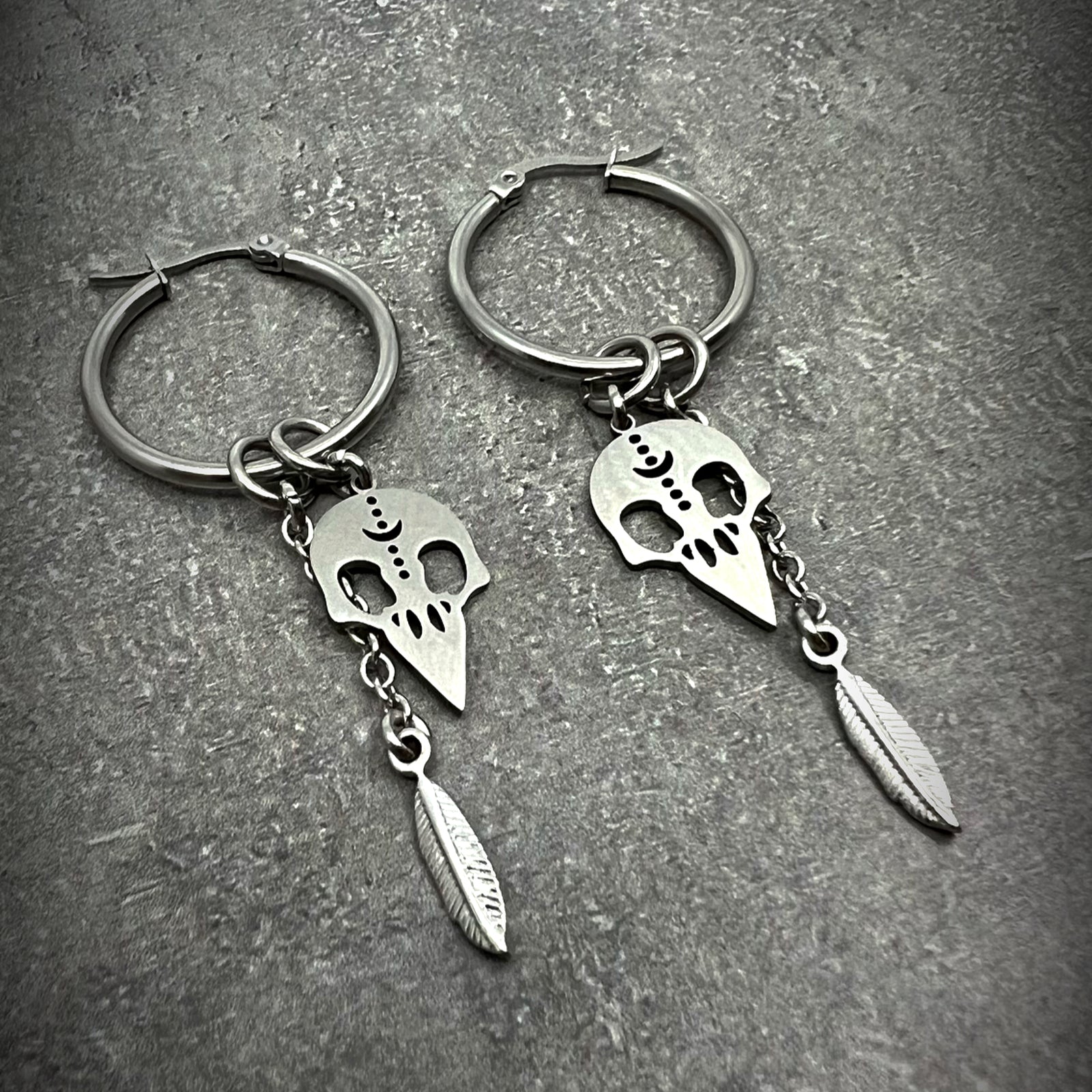 All-silver stainless steel hoop earrings featuring skull and feather charms, sculptural lightweight design — Lawless Jewellery.