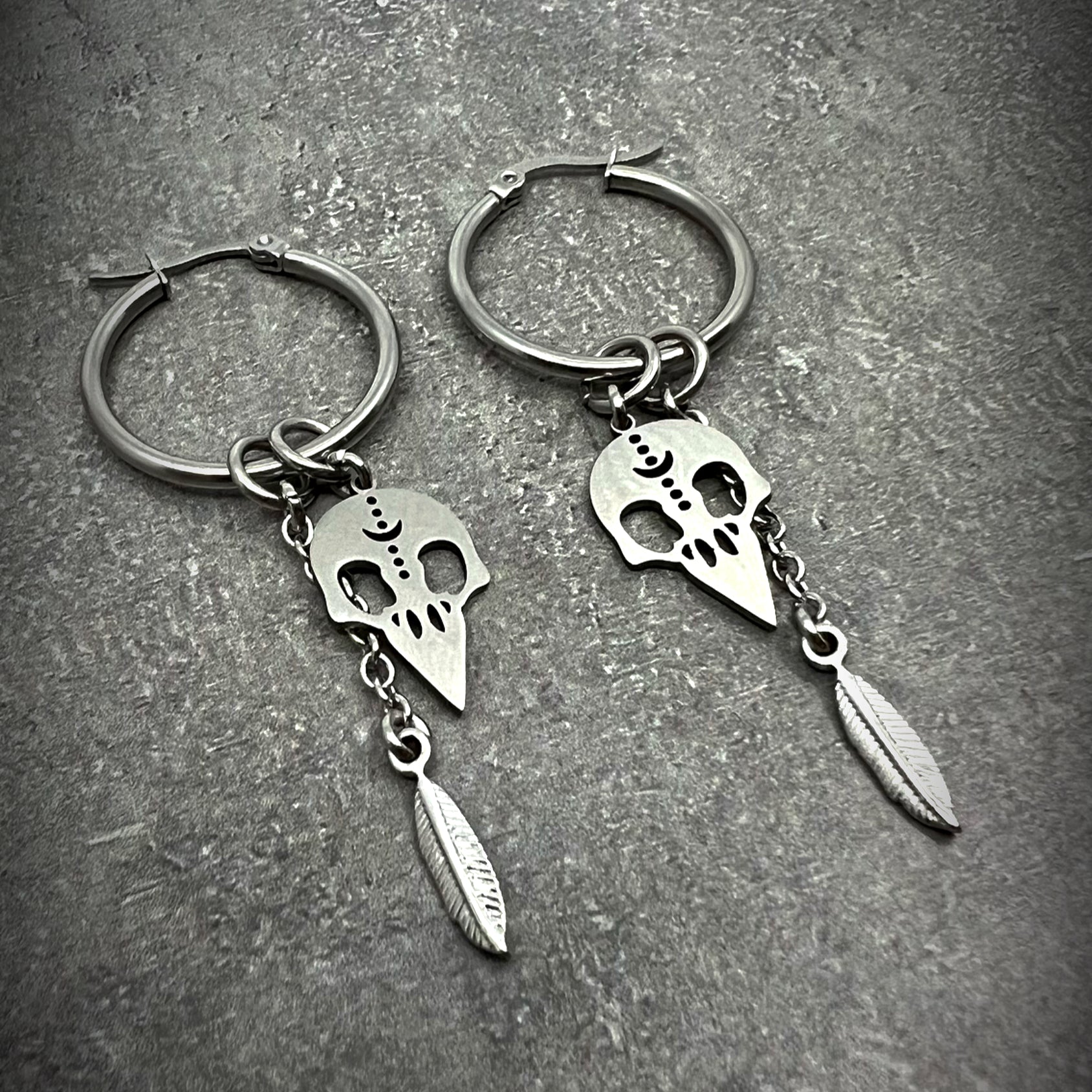 All-silver stainless steel hoop earrings featuring skull and feather charms, sculptural lightweight design — Lawless Jewellery.