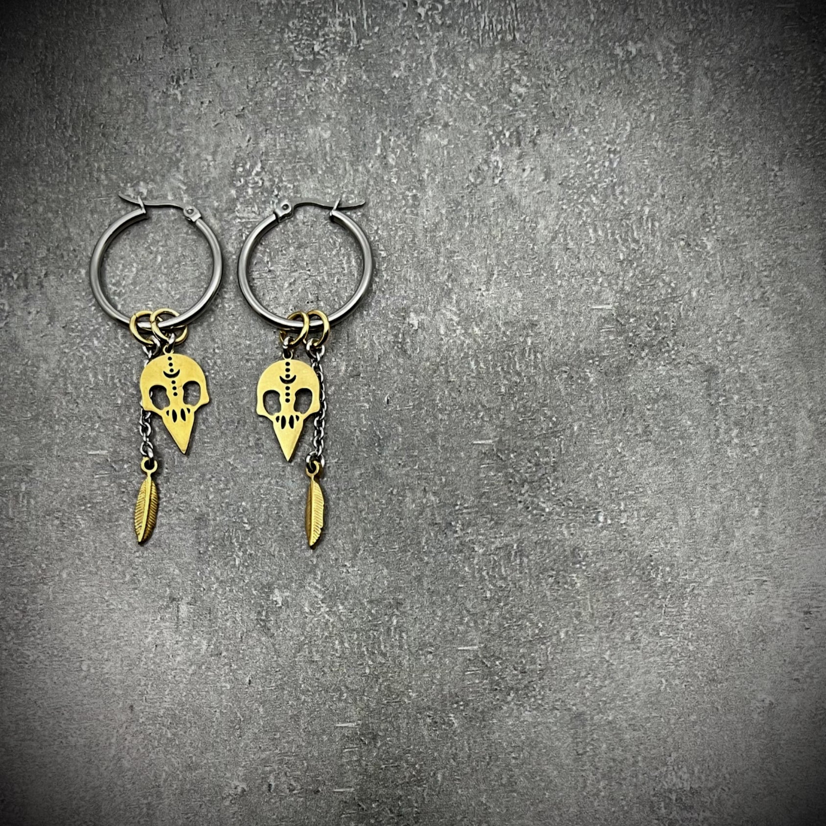 Silver stainless steel hoop earrings featuring gold skull and feather charms, lightweight dual-tone design — Lawless Jewellery.
