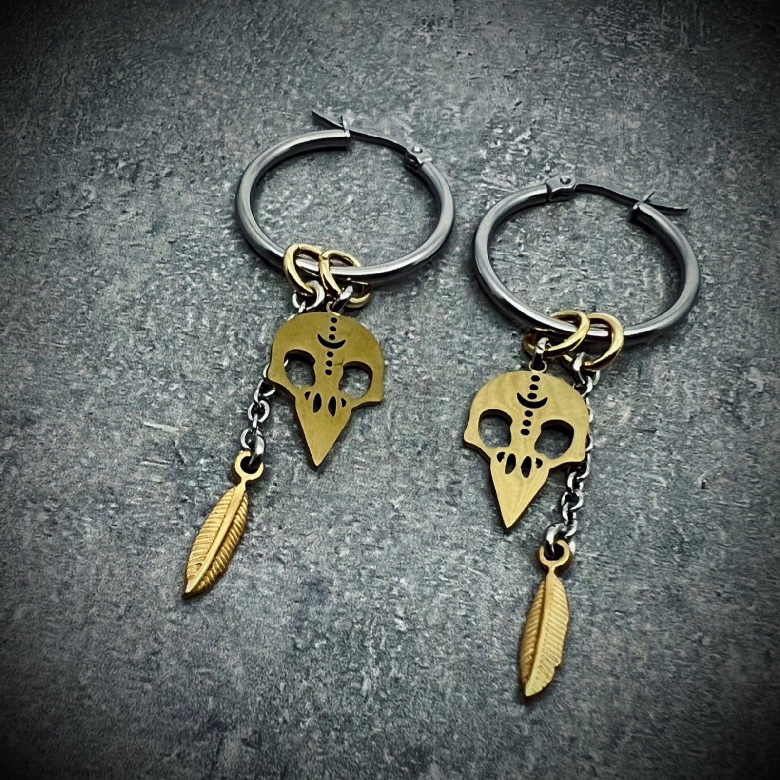 Silver stainless steel hoop earrings featuring gold skull and feather charms, lightweight dual-tone design — Lawless Jewellery.