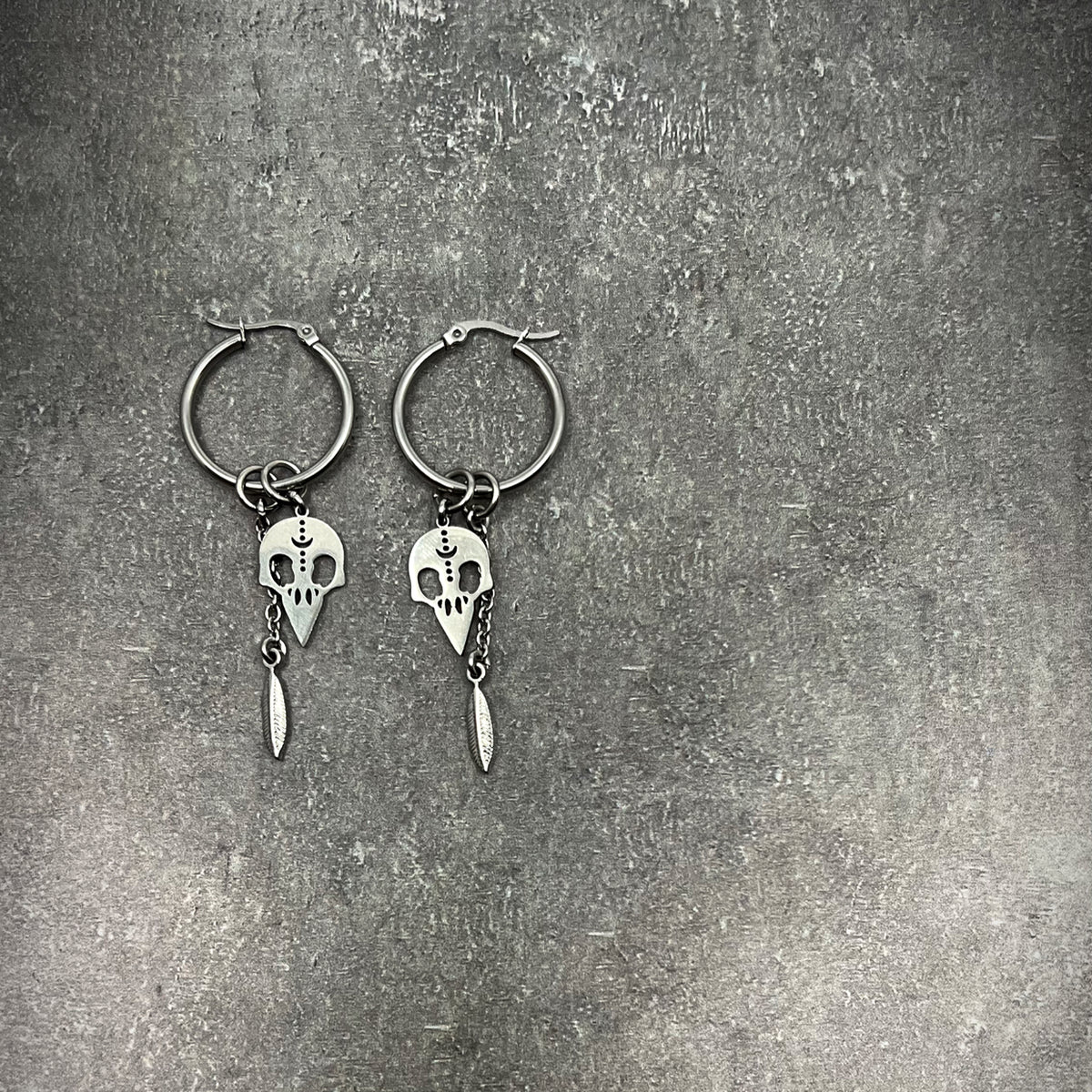 All-silver stainless steel hoop earrings featuring skull and feather charms, sculptural lightweight design — Lawless Jewellery.