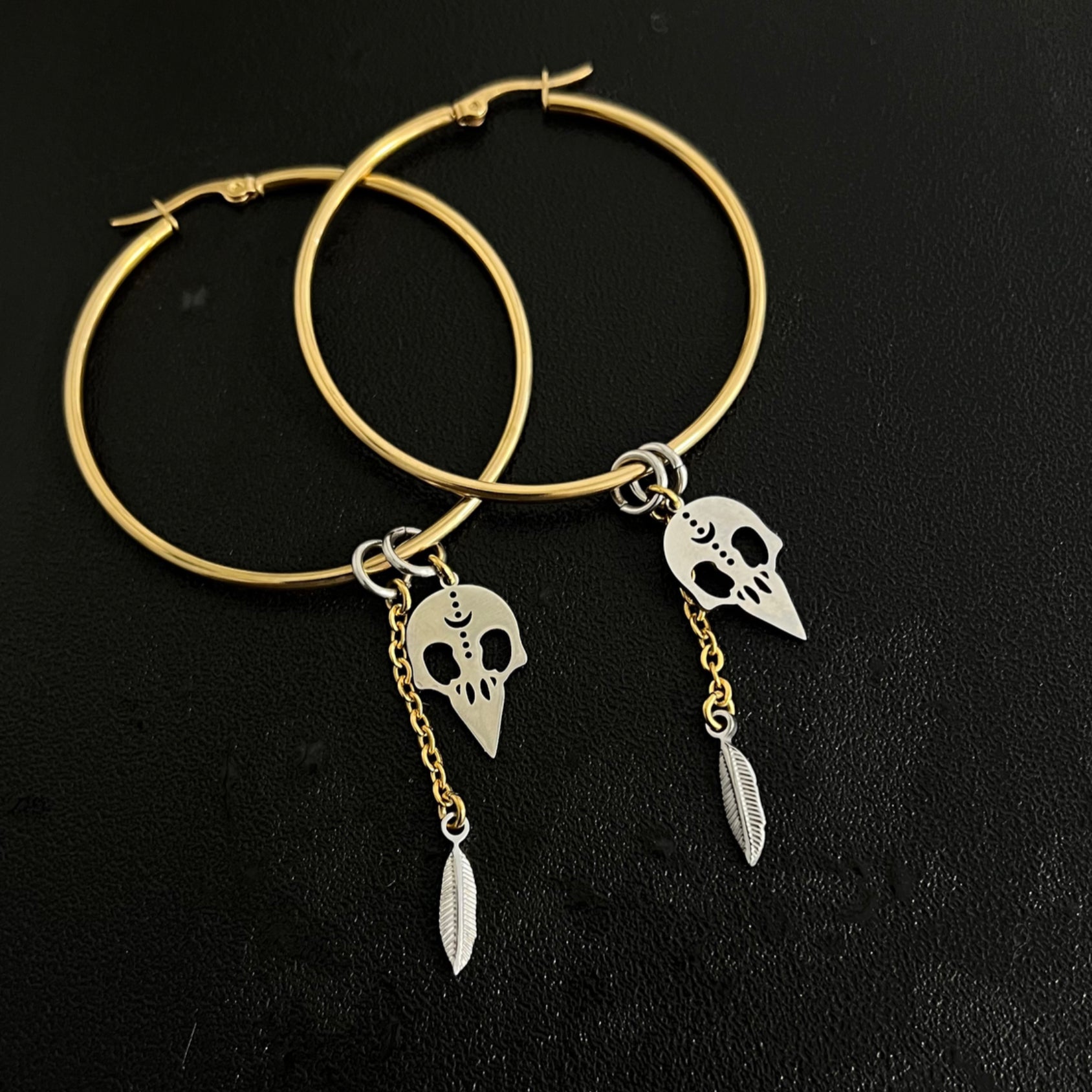 Oversized mixed-metal hoop earrings featuring gold skull and feather charms on silver stainless steel hoops — dual-tone statement design — Lawless Jewellery.