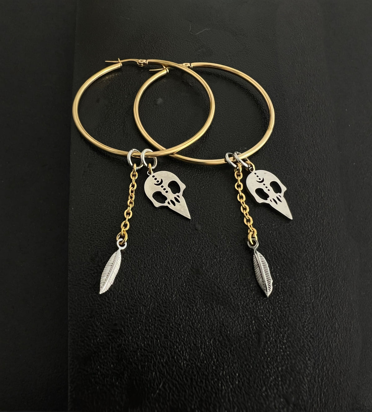 Oversized mixed-metal hoop earrings featuring gold skull and feather charms on silver stainless steel hoops — dual-tone statement design — Lawless Jewellery.