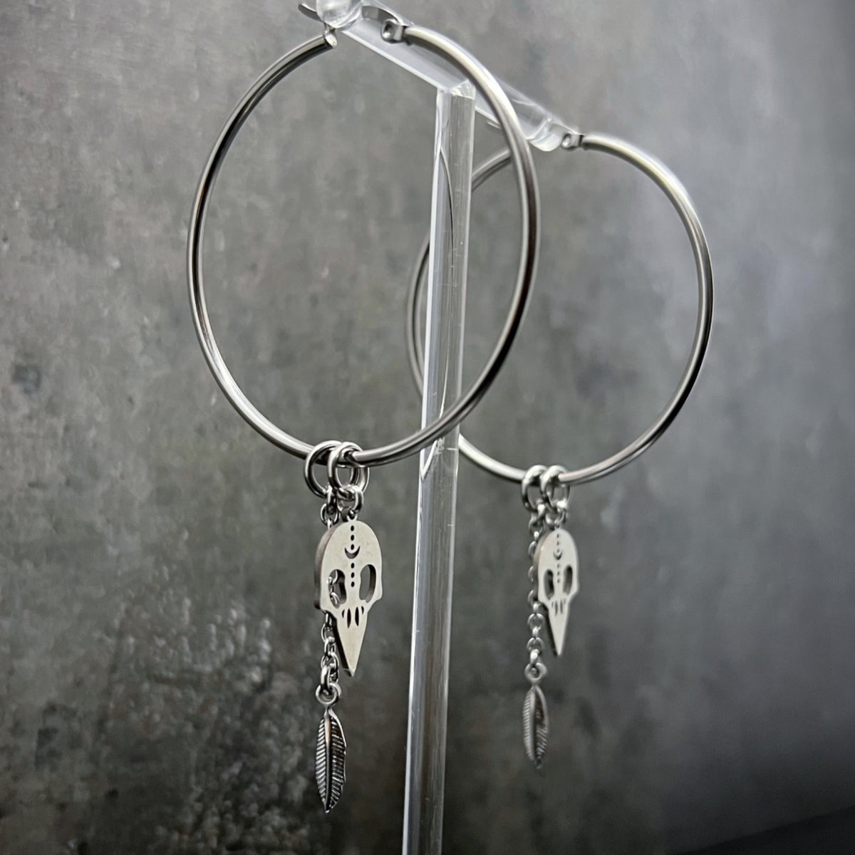 Oversized all-silver stainless steel hoop earrings featuring sculptural skull and feather charms — bold yet lightweight design — Lawless Jewellery.