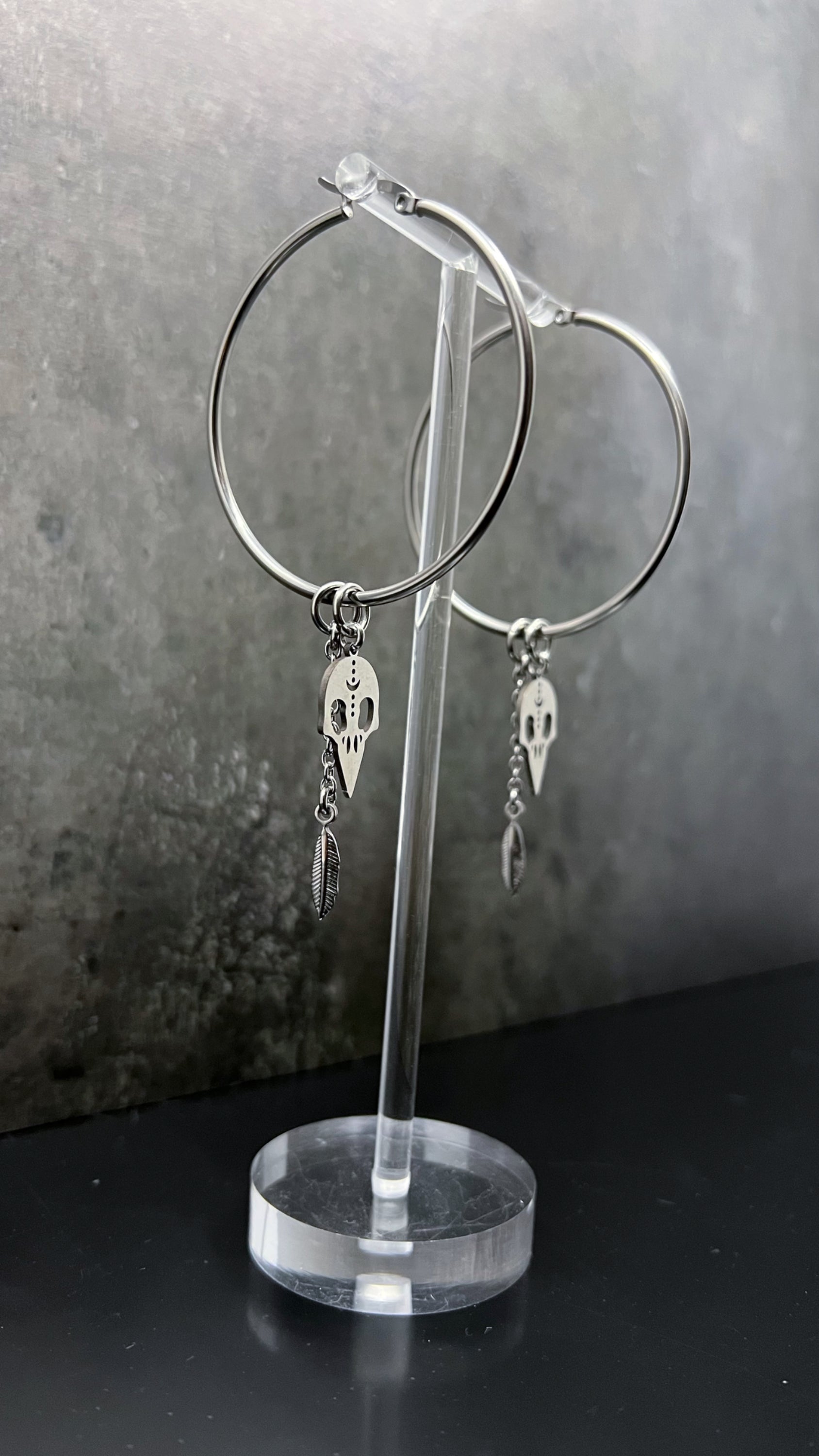 Oversized all-silver stainless steel hoop earrings featuring sculptural skull and feather charms — bold yet lightweight design — Lawless Jewellery.