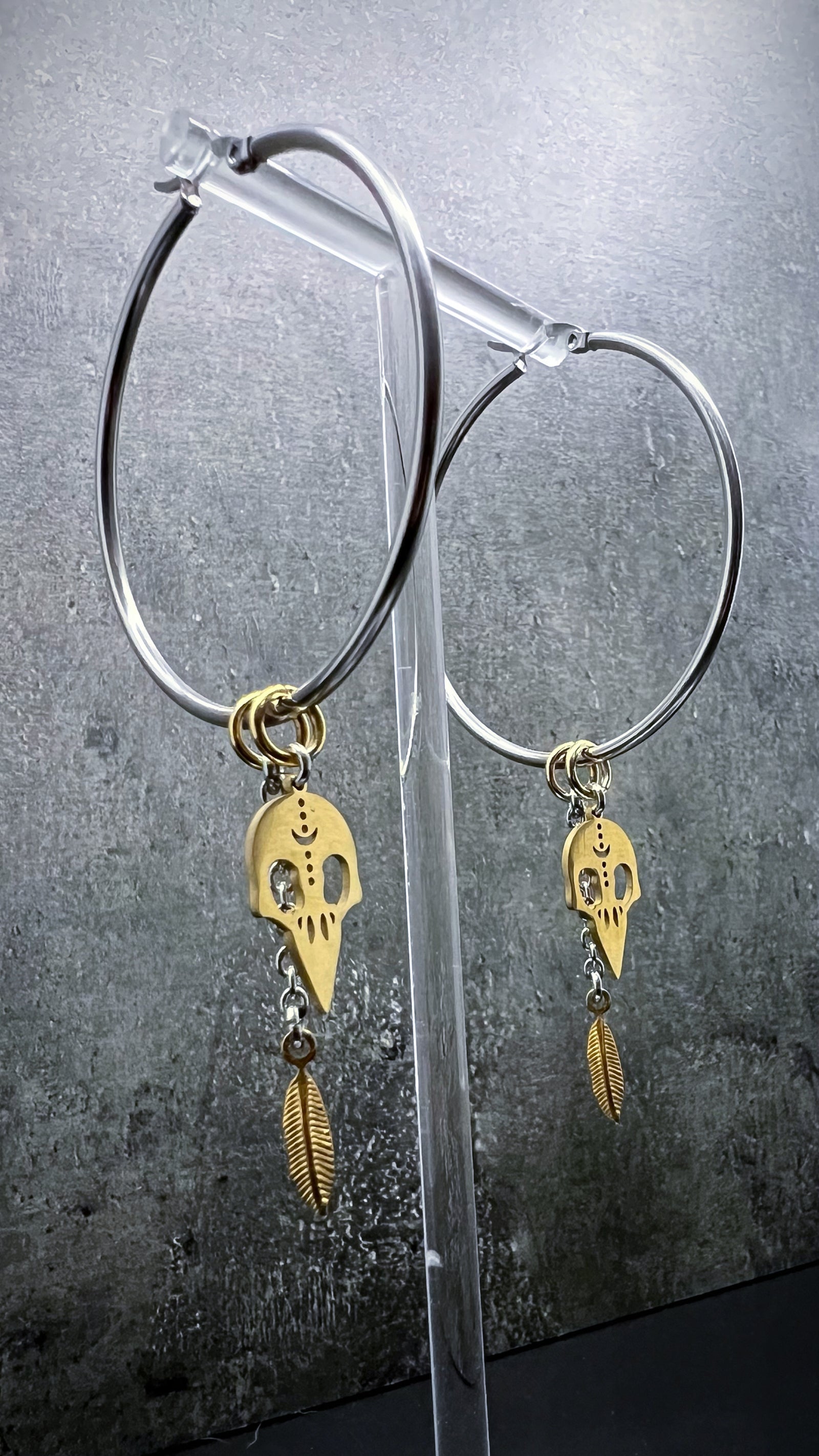 Oversized all-silver stainless steel hoop earrings featuring sculptural skull and feather charms — bold yet lightweight design — Lawless Jewellery.