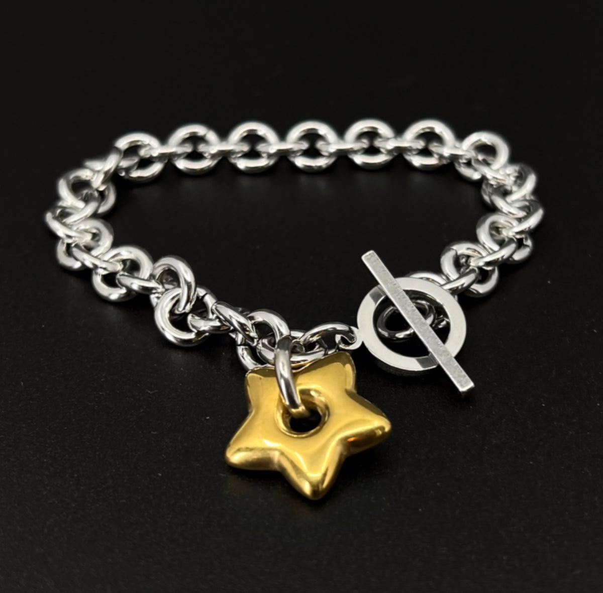 Mixed-metal stainless steel bracelet featuring an 18mm gold star charm on a silver chain with toggle clasp, photographed on a dark background.