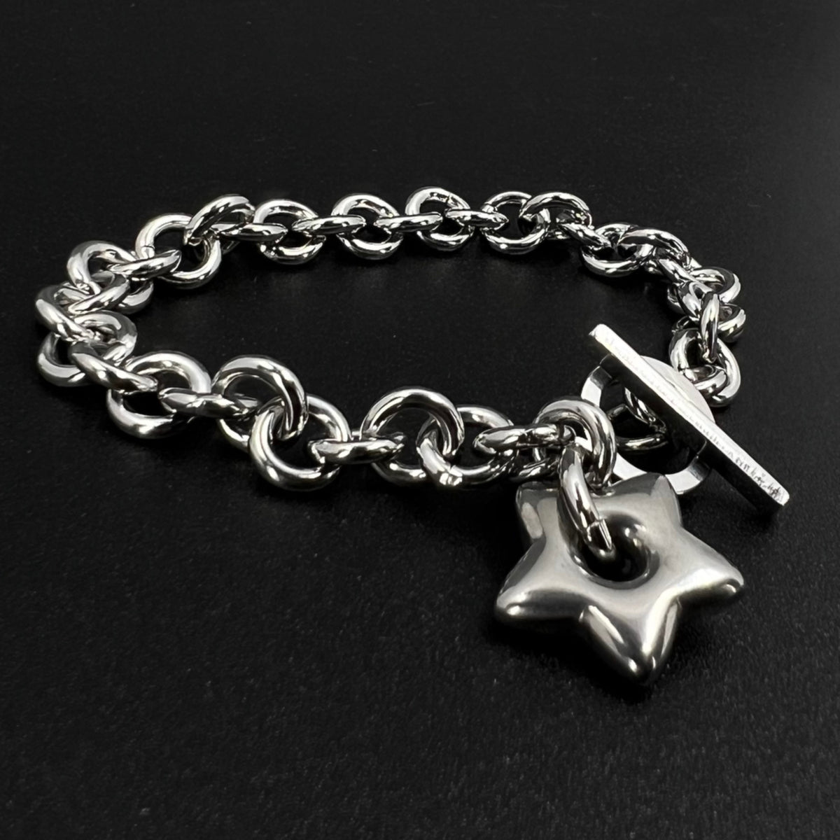 All-silver stainless steel bracelet featuring an 18mm star charm on a polished chain with toggle clasp, photographed on a dark background.