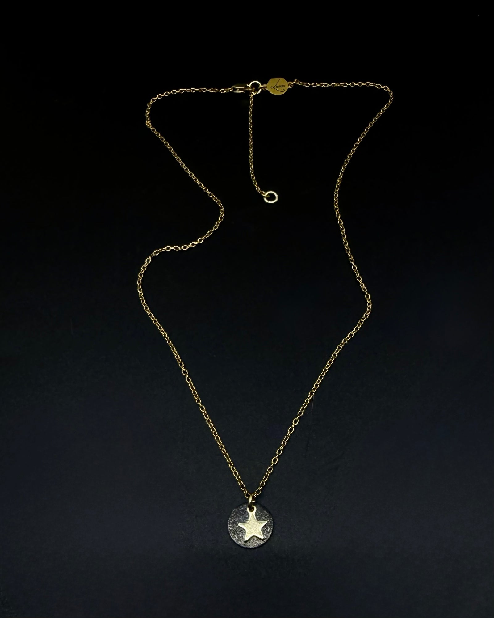 Black and gold Urania Signal Star Necklace featuring a textured black disc with a polished gold star charm on an adjustable stainless steel chain.