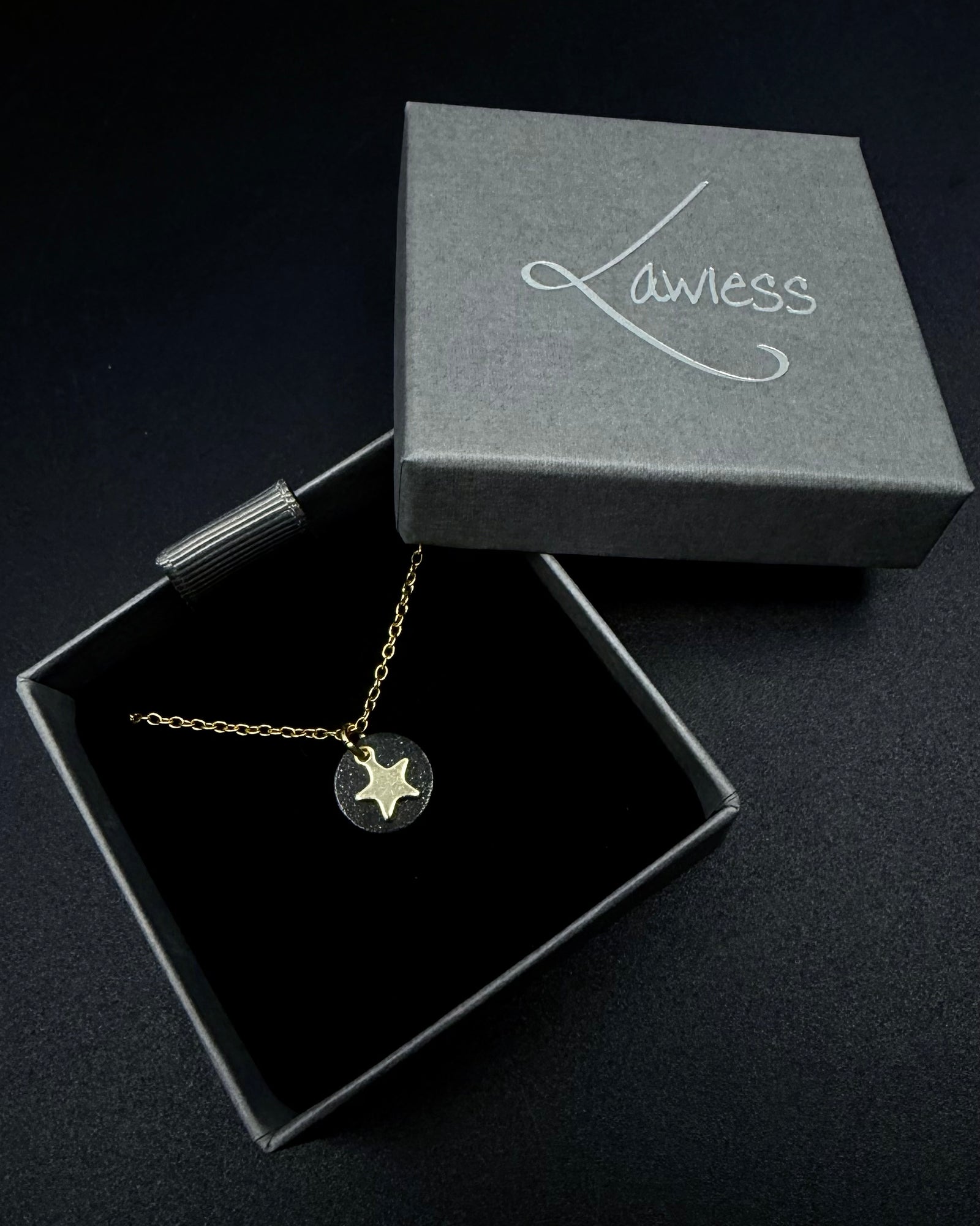 Black and gold Urania Signal Star Necklace featuring a textured black disc with a polished gold star charm on an adjustable stainless steel chain.