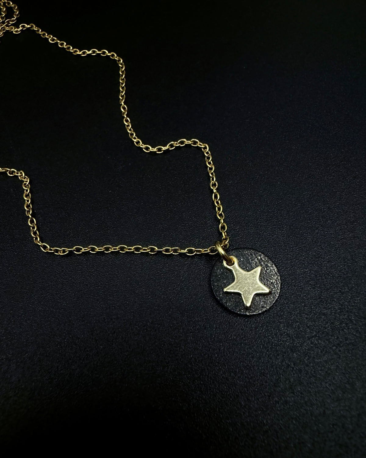Black and gold Urania Signal Star Necklace featuring a textured black disc with a polished gold star charm on an adjustable stainless steel chain.