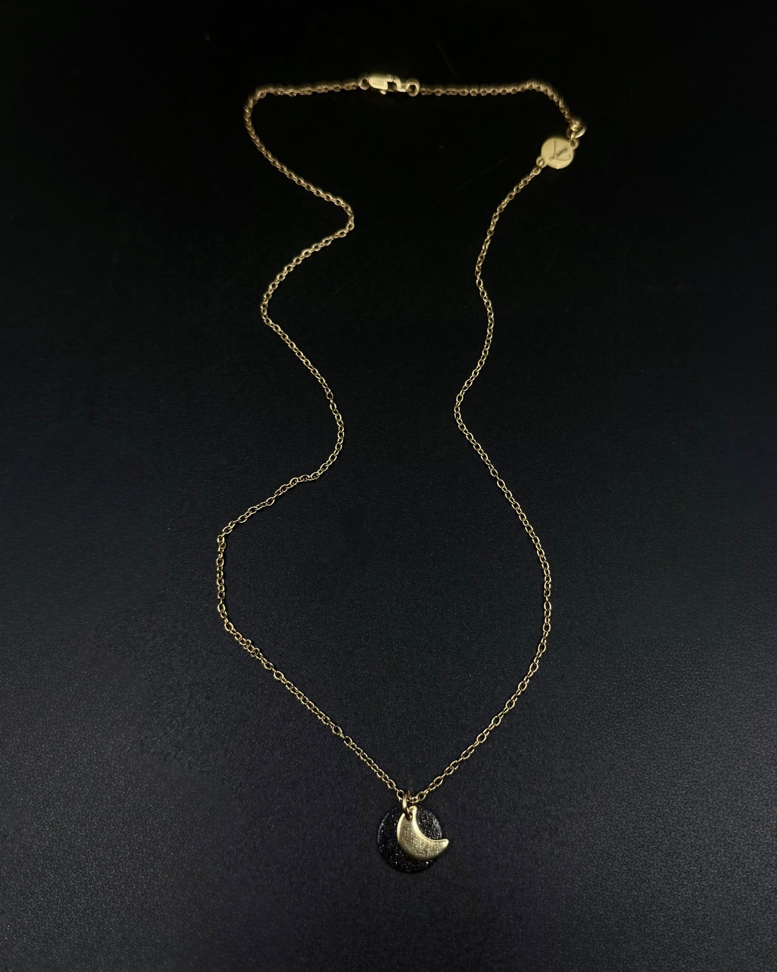Black and gold Urania Signal necklace with matte black disc and gold crescent moon pendant on adjustable stainless steel chain.